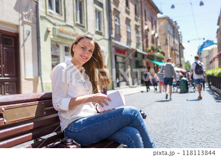 Young woman sitting on the bench in an old city and reading book on sunny day 118113844