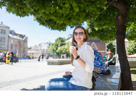 Young woman enjoying coffee outdoors under a tree in a sunny city square during the day Young woman enjoying coffee outdoors under a tree in a sunny city square during the day 118113847