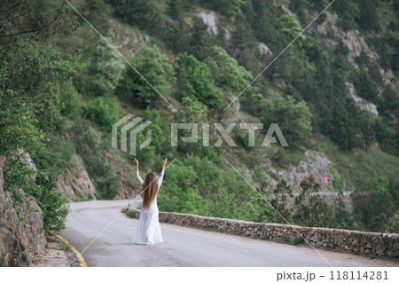 A woman is standing on a road, looking up at the sky. She is wearing a white dress and she is happy. The scene is set in a mountainous area, with trees and rocks in the background. 118114281