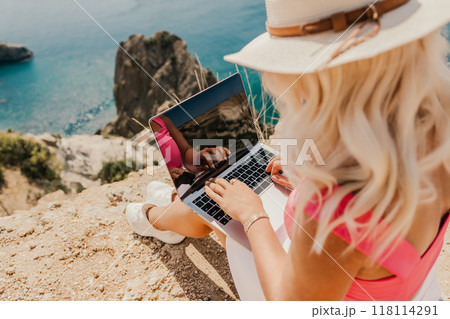A woman is sitting on a rock by the ocean, typing on her laptop. Concept of relaxation and leisure, as the woman enjoys the beautiful view of the water while working on her computer. A woman is sitting on a rock by the ocean, typing on her laptop. Concept of relaxation and leisure, as the woman enjoys the beautiful view of the water while working on her computer. 118114291