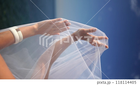 A woman is holding a white veil over her head. The woman is standing on a beach, and the ocean is visible in the background. A woman is holding a white veil over her head. The woman is standing on a beach, and the ocean is visible in the background. 118114405