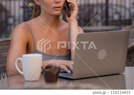 Woman Laptop Phone Call Cafe - A woman sits at a cafe table with a laptop and a phone, talking on the phone while working. Woman Laptop Phone Call Cafe - A woman sits at a cafe table with a laptop and a phone, talking on the phone while working. 118114455