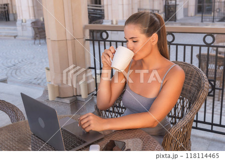 Woman Coffee Laptop - Woman sits outdoors, drinking coffee and working on her laptop. Woman Coffee Laptop - Woman sits outdoors, drinking coffee and working on her laptop. 118114465