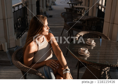 Woman Coffee Cafe - A young woman sits at a cafe table with a cup of coffee, enjoying the afternoon sun. 118115416