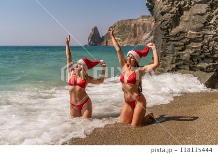 Women Santa hats ocean play. Seaside, beach daytime, enjoying beach fun. Two women in red swimsuits and Santa hats are enjoying themselves in the ocean, kneeling in the waves and raising their hands 118115444