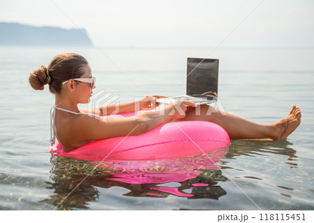 A woman is sitting in a pink inflatable raft on a lake, using a laptop. Concept of relaxation and leisure, as the woman is enjoying her time outdoors while working on her laptop. 118115451