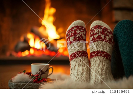 Feet in woollen socks by the Christmas fireplace. Woman relaxes by warm fire with a cup of hot drink and warming up her feet in woollen socks. Close up on feet. Winter and Christmas holidays concept. 118126491