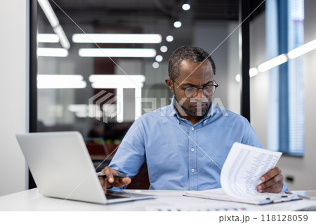 Business professional concentrating on reviewing documents at desk with laptop in modern office. Man appears focused and engaged, highlighting concept of productivity and attention to detail. Business professional concentrating on reviewing documents at desk with laptop in modern office. Man appears focused and engaged, highlighting concept of productivity and attention to detail. 118128059