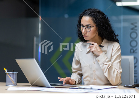 Hispanic businesswoman in formal shirt working on laptop, focused and serious in office setting. Concentrating on important tasks, holding pen, demonstrating professional mindset. Hispanic businesswoman in formal shirt working on laptop, focused and serious in office setting. Concentrating on important tasks, holding pen, demonstrating professional mindset. 118128671