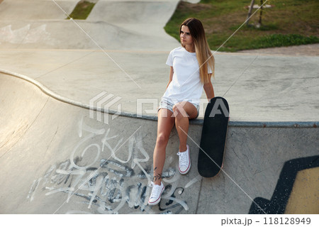 Attractive girl with a tattoo in a white t-shirt sitting on a ramp in skate park with a skateboard. 118128949