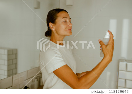 A woman in a beautifully bright bathroom is applying a skincare product with grace and care, 118129645