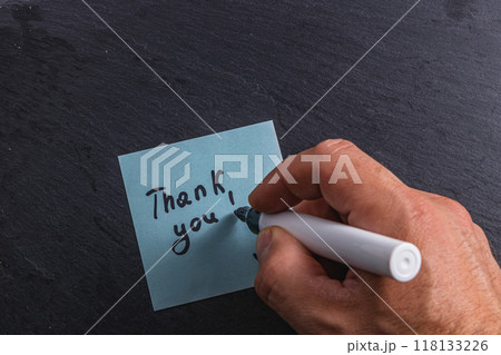 A man writes the word thank you on a blue piece of paper on a table A man writes the word thank you on a blue piece of paper on a table 118133226