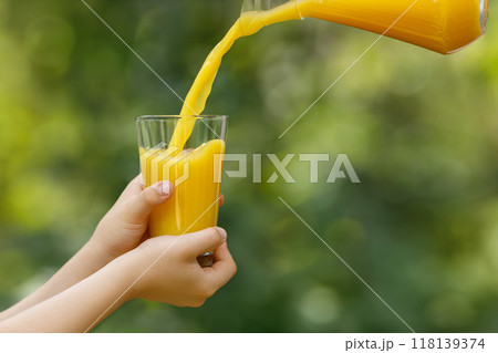 child hands holding glass and orange juice pouring from jug with green garden on the background 118139374