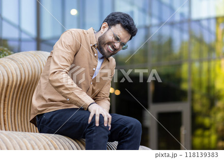 Man wearing glasses and casual clothing sitting on modern design wooden bench holding knee. Expression showing discomfort and pain while seated outdoors. Captured in natural light in urban setting. Man wearing glasses and casual clothing sitting on modern design wooden bench holding knee. Expression showing discomfort and pain while seated outdoors. Captured in natural light in urban setting. 118139383