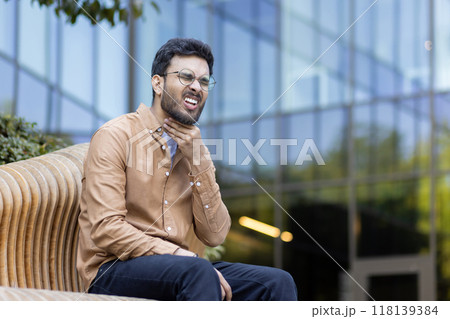 Young man sitting outdoors holding throat with pained expression. Wearing glasses and casual brown shirt with glass building in background. Concepts include pain, discomfort, health concern. Young man sitting outdoors holding throat with pained expression. Wearing glasses and casual brown shirt with glass building in background. Concepts include pain, discomfort, health concern. 118139384