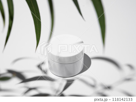 Face and body cream in a white jar on a light background with leaves and hard shadow. Face and body cream in a white jar on a light background with leaves and hard shadow. 118142133