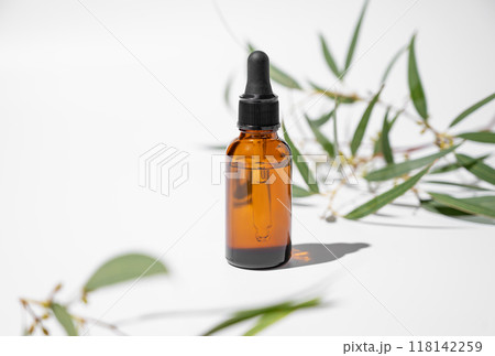 Face and body serum in bottle on a white background with green leaves and shadow. 118142259