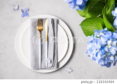 Festive table setting with blue hydrangea flowers and white plates on a light background.  118142484