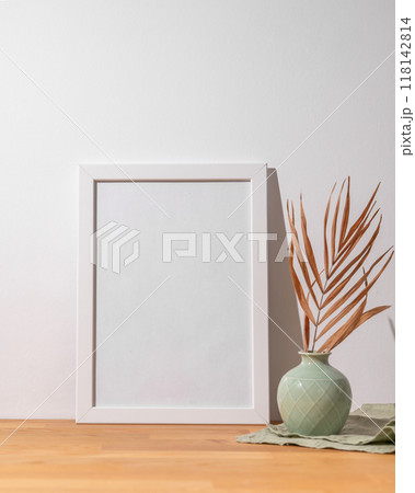 A white frame with a blank canvas against a light wall and on a wooden tabletop with vase. 118142814