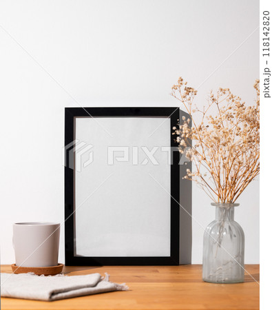A black frame with a blank canvas against a light wall and on a wooden tabletop with vase and cup. A black frame with a blank canvas against a light wall and on a wooden tabletop with vase and cup. 118142820
