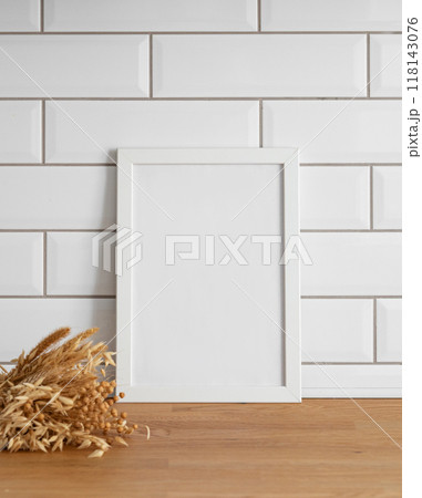 A frame with a blank canvas against a white tile wall and on a wooden tabletop with dry bouquet. A frame with a blank canvas against a white tile wall and on a wooden tabletop with dry bouquet. 118143076