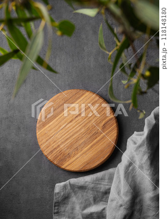 Wooden empty round board with green leaves and napkin on a dark gray background. 118148180