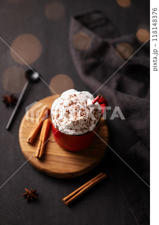 Red mug with hot chocolate or cocoa with whipped cream on a wooden stand with cinnamon sticks  118148376
