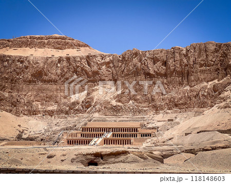 photo shows an ancient temple nestled against towering rocky cliffs in the desert. The warm tones of the stone contrast with the deep blue sky, emphasizing the harmony between architecture and nature. 118148603