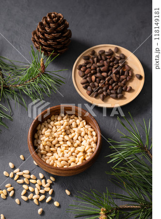Pine nuts in a bowl and a scattered on a dark background with branches of pine needles and cone. 118149181