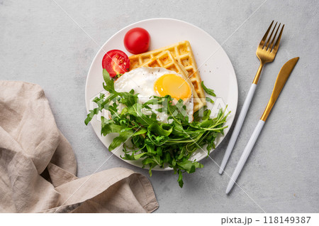 Waffles with fried egg, cherry tomatoes and arugula on a white plate on a gray texture background Waffles with fried egg, cherry tomatoes and arugula on a white plate on a gray texture background 118149387