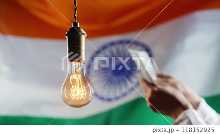 Glowing Edison light bulb on Indian flag background. Woman counting rupee INR money. Electricity price, bills, energy crisis, raising tariffs. 118152925