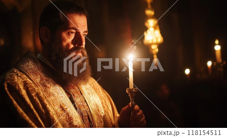 Close up portrait of an Orthodox priest with a long beard wearing an ornate golden robe, holding a lit candle in a dimly lit church setting 118154511