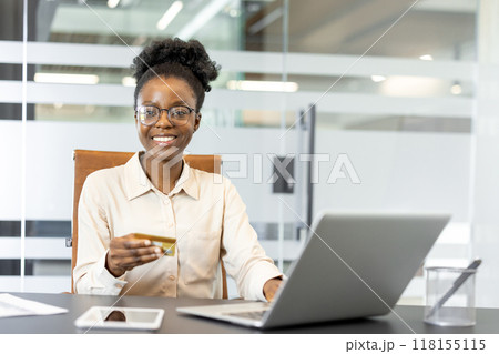 Confident African American businesswoman using laptop while holding credit card, symbolizing online shopping or e-commerce in professional office environment. Smartphone on table adds technological 118155115