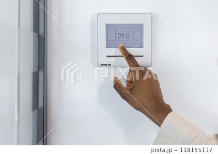Close-up of hand touching digital thermostat screen showing 26 degrees Celsius. Thermostat mounted on office wall manages indoor climate for optimal comfort and energy efficiency. 118155117