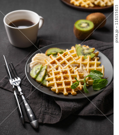 A freshly prepared waffles with kiwi and banana slices on a gray plate with a cup of coffee  118155815