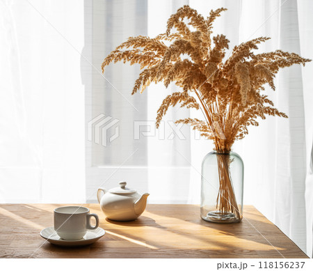 A cup of tea, teapot and dry bouquet flowers on the wooden table of a window with morning sunlight. A cup of tea, teapot and dry bouquet flowers on the wooden table of a window with morning sunlight. 118156237