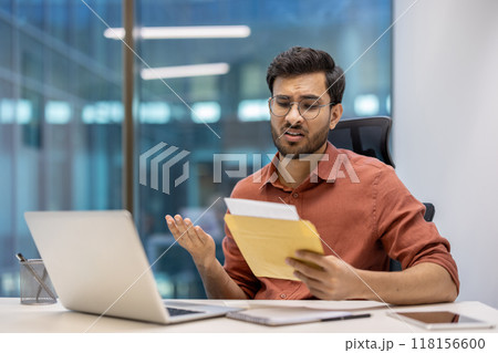 Young professional in office environment appearing frustrated after receiving bad news in postal envelope. Seated at desk with laptop, phone, and documents. Young professional in office environment appearing frustrated after receiving bad news in postal envelope. Seated at desk with laptop, phone, and documents. 118156600