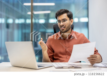 Office worker with glasses showing concern over financial report at desk with laptop. Business professional analyzing documents, displaying worry. Concept stress and dissatisfaction in financial work 118156622