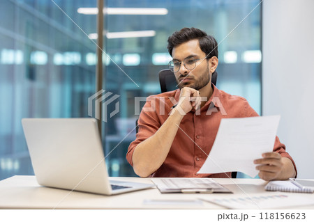 Serious businessman thinking while reviewing document at office desk. Professional wearing glasses focused on paperwork with laptop in front. Calm environment reflects dedication, concentration 118156623