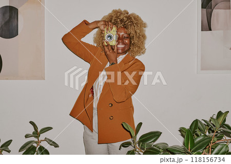 Smiling curly haired woman holding yellow camera and surrounded by ficuses while imitating photographer Smiling curly haired woman holding yellow camera and surrounded by ficuses while imitating photographer 118156734