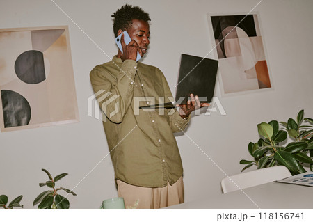 Dutch angle shot of busy young influencer holding laptop while speaking on phone Dutch angle shot of busy young influencer holding laptop while speaking on phone 118156741