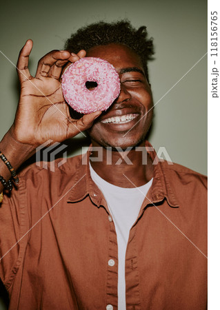 Young man holding pastel pink doughnut to hide his eye while posing for photo Young man holding pastel pink doughnut to hide his eye while posing for photo 118156765