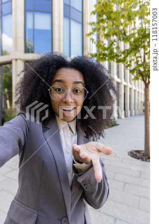 Confident African American businesswoman making video call using phone outdoors. Professional attire, smiling expression. Urban environment, modern architecture, connection, communication 118157003