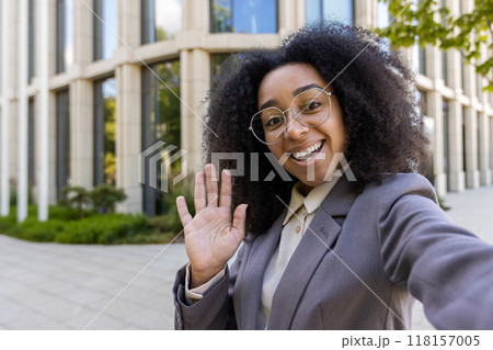 Confident African American business woman waving during phone video call. Standing in front of contemporary office building. Professional and friendly demeanor enhances business communication. Confident African American business woman waving during phone video call. Standing in front of contemporary office building. Professional and friendly demeanor enhances business communication. 118157005