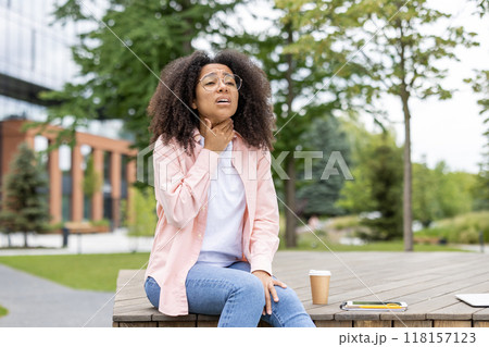 Woman sitting outside holding throat, expressing discomfort, possibly from soreness or pain. She is seated on wooden bench with phone and coffee cup visible. Urban park setting with greenery around. Woman sitting outside holding throat, expressing discomfort, possibly from soreness or pain. She is seated on wooden bench with phone and coffee cup visible. Urban park setting with greenery around. 118157123