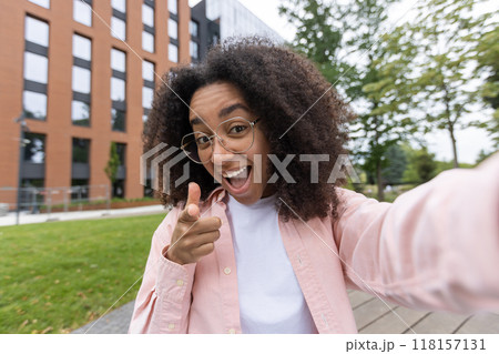 Smiling woman taking selfie outdoors with cheerful expression pointing at camera. Urban setting with trees and building adds vibrant atmosphere. Captures joy, confidence, and connection Smiling woman taking selfie outdoors with cheerful expression pointing at camera. Urban setting with trees and building adds vibrant atmosphere. Captures joy, confidence, and connection 118157131