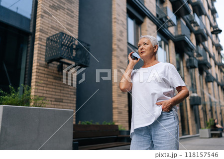 Low-angle view of senior Caucasian female focused talking on smartphone standing against urban backdrop with soft lighting. Happy older woman smiling talking on smartphone with family outside. Low-angle view of senior Caucasian female focused talking on smartphone standing against urban backdrop with soft lighting. Happy older woman smiling talking on smartphone with family outside. 118157546
