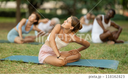 Girl performing stretches in lotus position during group yoga session in park Girl performing stretches in lotus position during group yoga session in park 118162823