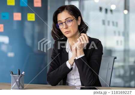 Professional woman wearing glasses and business suit sitting at office desk looking at camera. Confident expression and composed demeanor, ready for work or business meeting. Professional woman wearing glasses and business suit sitting at office desk looking at camera. Confident expression and composed demeanor, ready for work or business meeting. 118169227
