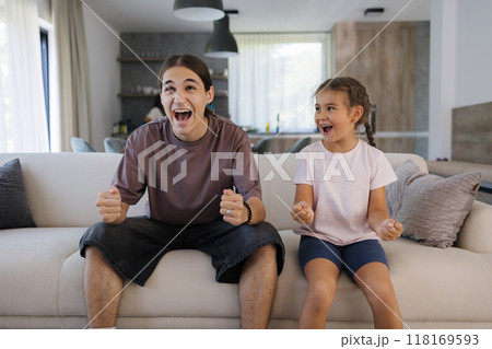Brother and sister watching TV and reacts emotionally to the events in the TV show. Teenage boy and little girl sitting on couch at home.  118169593
